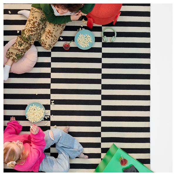 Three kids in different outfits on a striped rug snacking popcorn from blue plates. Headphones and remote nearby.