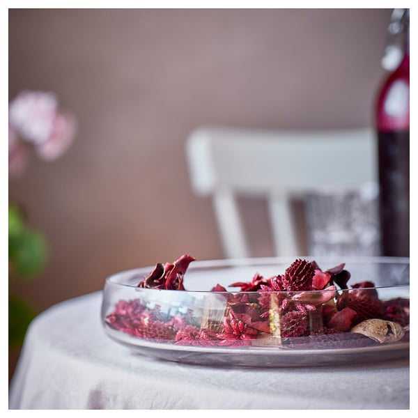 Clear bowl with red potpourri, including leaves and pinecones, on a white table. Soft focus background.