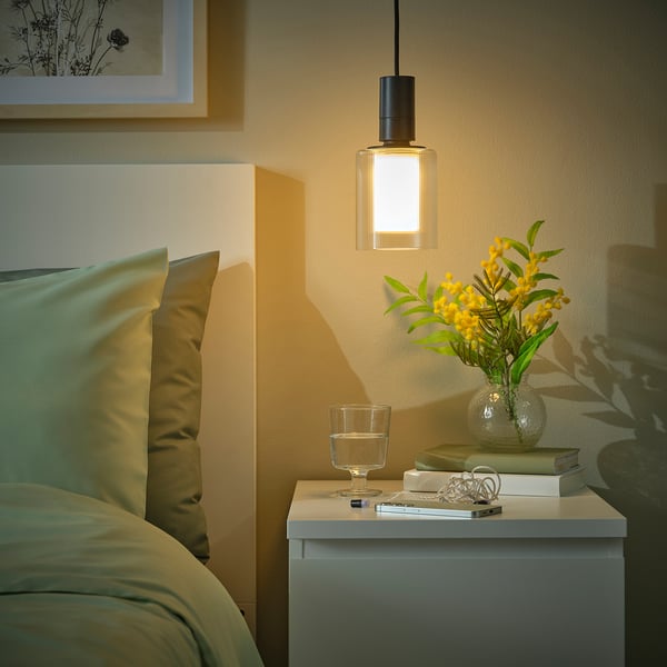 A cosy bedroom with a hanging SUNNEBY lamp emitting warm light, a bedside table with books, a glass, and flowers, and a bed with green pillows.