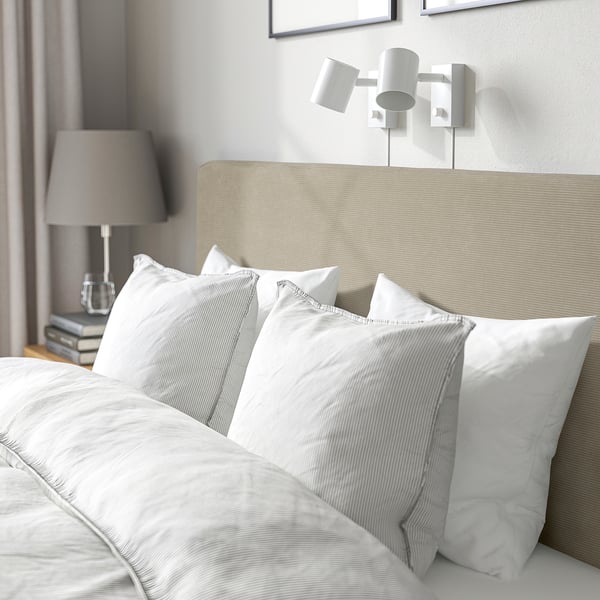 A neatly made bed with a soft, beige headboard, four fluffy white pillows, and a side table lamp. The beds cover is removable and washable.