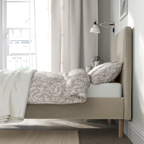 Cosy bed with beige headboard, floral bedding, and wall lamp. Near large, curtained windows.