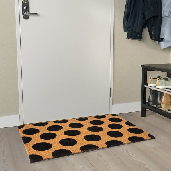 Black and tan welcome mat with dots sits by a white door on light wood floor.