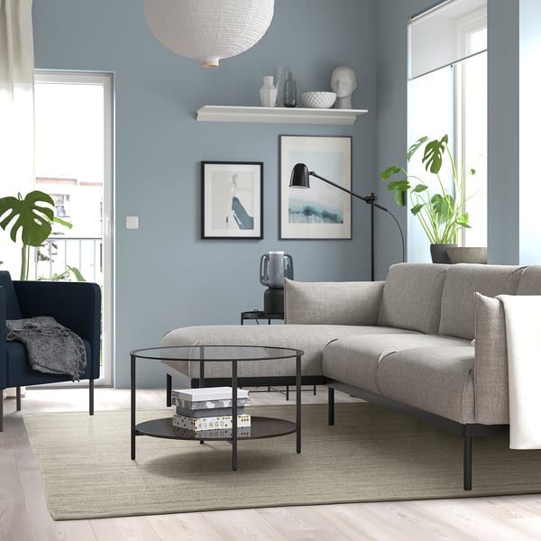 Modern living room with grey TIDTABELL rug, coffee table, grey sofa, dark blue armchair, art on light blue walls, window with plant.
