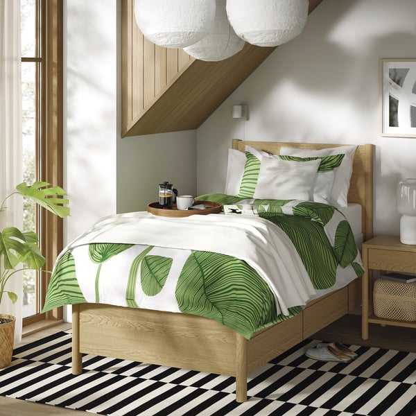Bedroom with TONSTAD light wood bed, green leaf print linens, black-white rug, large plant, hanging lamps, white walls.
