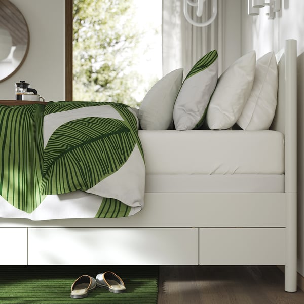 A serene, white bedroom scene. Green and white bedding with palm leaves, white pillows. A pair of black sandals under a white bed.