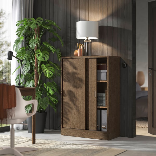 TONSTAD wooden storage cabinet with sliding doors, holding office supplies. Adjustable shelves, beside a plant, chair, and desk.