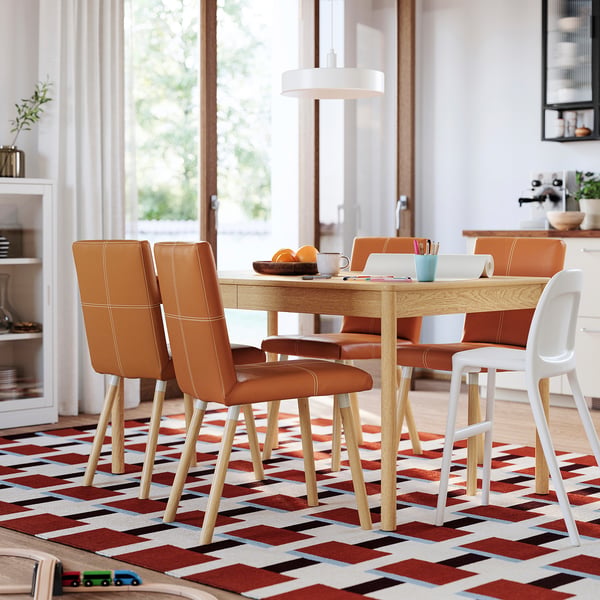 Bright dining room with orange leather chairs, white stool, wooden table, colourful rug, and large window.