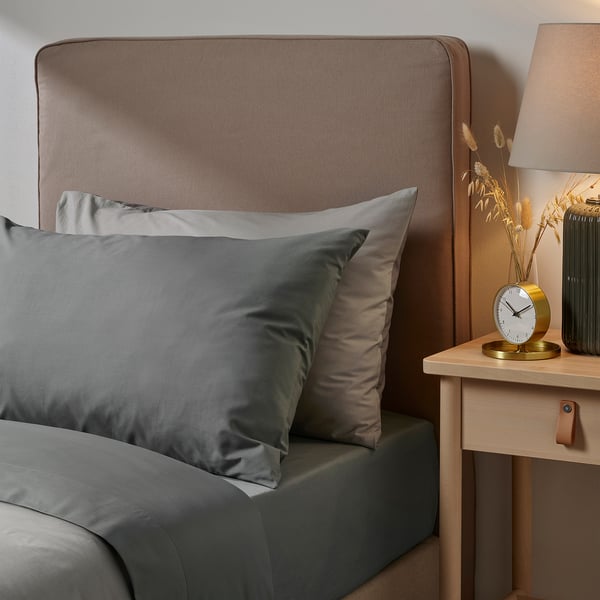 Modern bedroom scene with grey ULLVIDE bedding on large bed; wooden bedside table holds gold clock, green vase with dried flowers, and brown lamp.