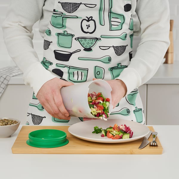 Person pouring salad from a white UPPFYLLD salad shaker onto a plate with cutlery on a wooden board.