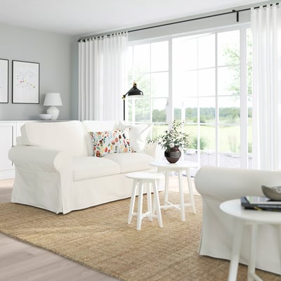 Bright white sofa, large, deep-seat, with colourful pillows. Situated near large windows, rolls heavy jute rug, surrounded by minimalist decor.