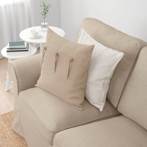 Beige sofa with reversible cushions, two throw pillows, side table with books, vase, and cup in bright, cosy space.
