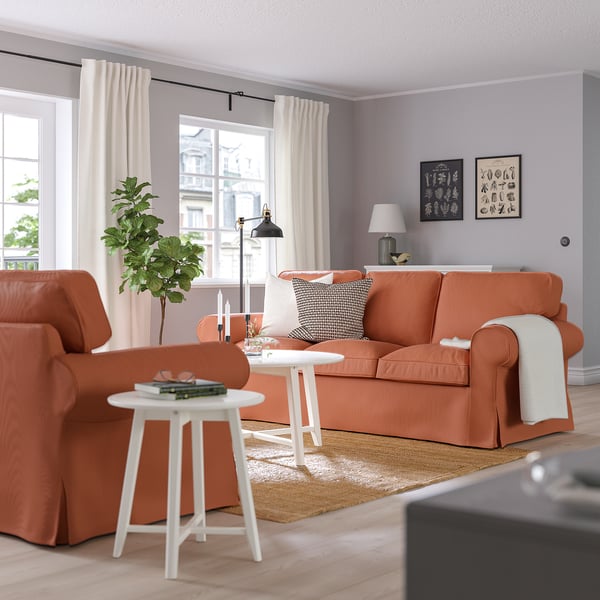 Bright, orange UPPLAND sofa dominates living room, flanked by white coffee tables. Accented with decor and plants, set against lavender walls.