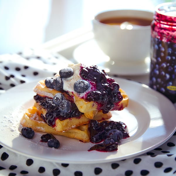 White plate with golden waffles, blueberries, cream, and dark sauce. Next to a white coffee cup and a jar of blueberries.
