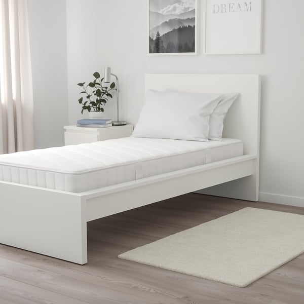 Modern single bed with white mattress & pillows, small bedside table with plant & lamp, light-colored rug, minimalist decor, wall art, curtains.