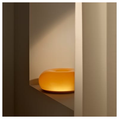 A modern, orange glass lamp sits on a shelf, emitting warm light. Its unique donut shape and sleek design make it an attractive decor piece.