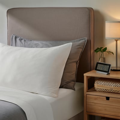 Grey bedding set with white sheets & pillows on mattress next to wooden bedside table with clock, lamp, and plant. Unique brown patterned headboard.
