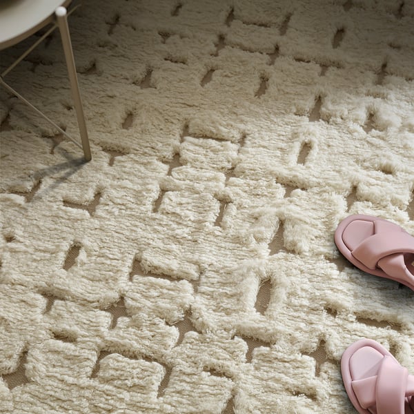 Cosy, cream-colored rug with geometric pattern and varied pile heights. Pink sandals rest on it, suggesting comfort and warmth.