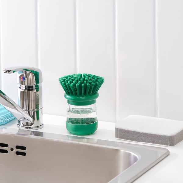 A green dish brush stands near a sink tap, filled with soap for easy dish cleaning.