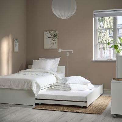 White bed frame with trundle, white linens, minimalist design. Small rug, lamp, greenery view.