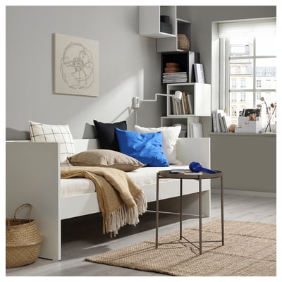 White daybed with blue pillows, side table, bookshelf, and window.