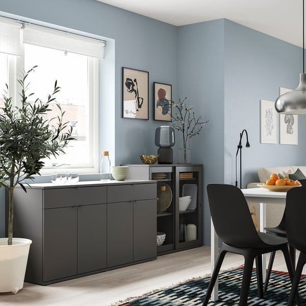 Modern living room with grey sideboard, black chairs, wall art, plant, and dining table with fruits.