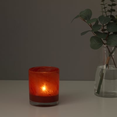 Lit orange glass candle holder on table, emitting warm light, next to clear vase with greenery.
